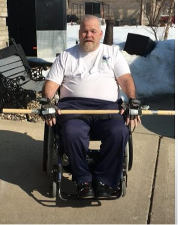 Brian sitting in wheelchair, white short sleeve t-shirt, outside in sun, snow pile behind, with exercise pole